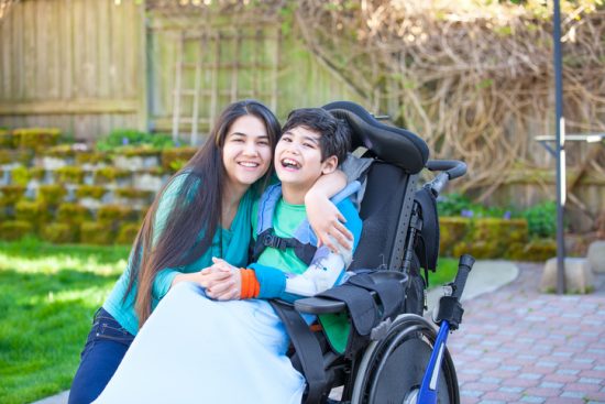 Smiling teenage girl hugging disabled nine year old brother in wheelchair outdoors